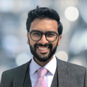 Headshot of Samir Janmohamed smiling, wearing glasses and a suit with a pink tie