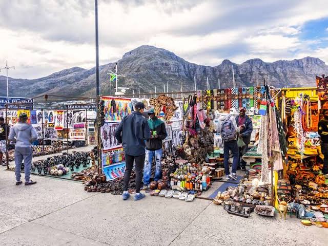 Bay Harbour Market: Visitors browse colorful stalls selling local crafts, artwork, and souvenirs at the Bay Harbour Market in Cape Town, with mountains in the background.