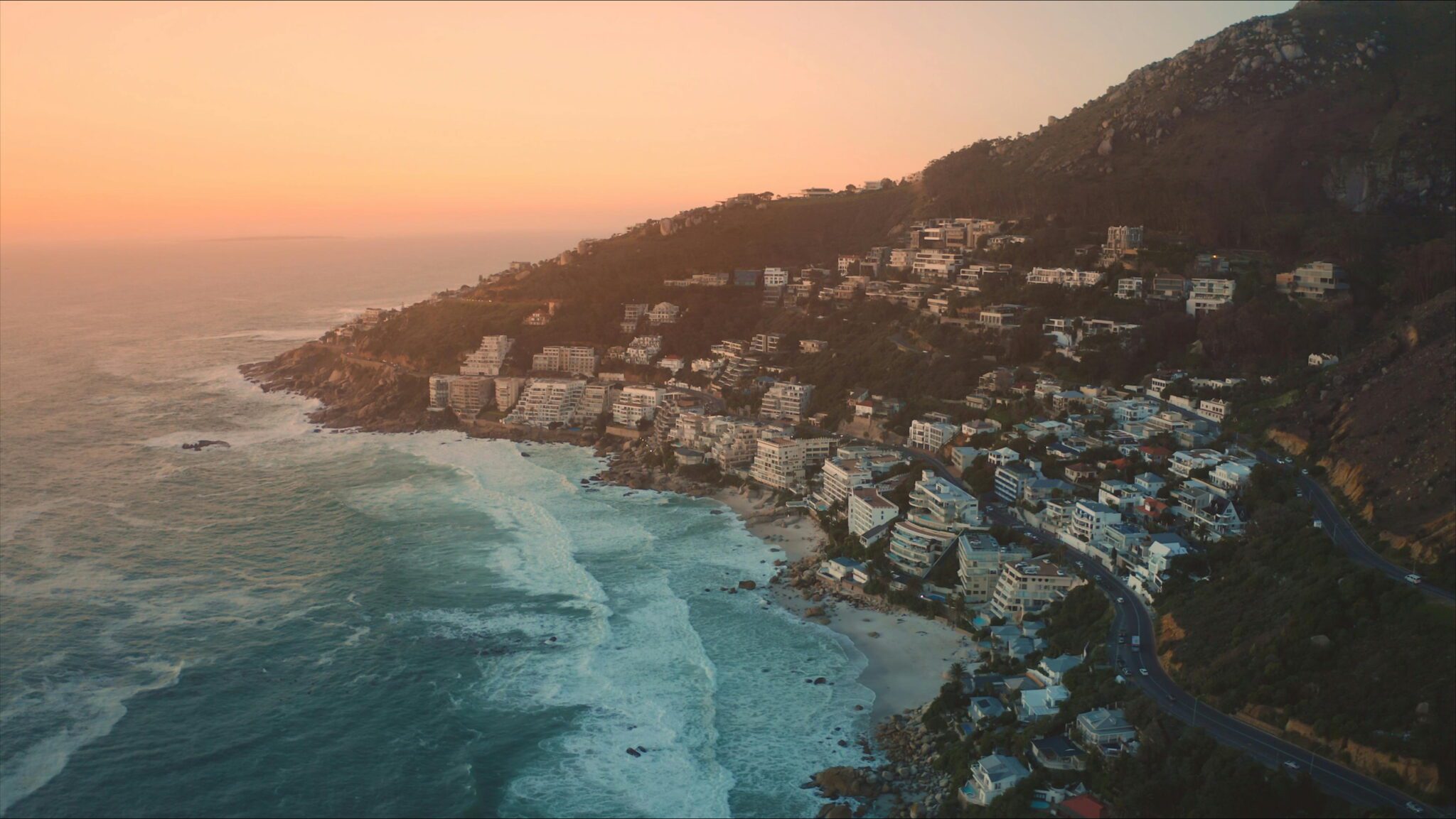 Clifton’s Fourth Beach: A scenic aerial view of Clifton’s Fourth Beach in Cape Town at sunset, showing waves crashing along the coastline, with terraced houses and buildings nestled on a steep hillside overlooking the ocean.