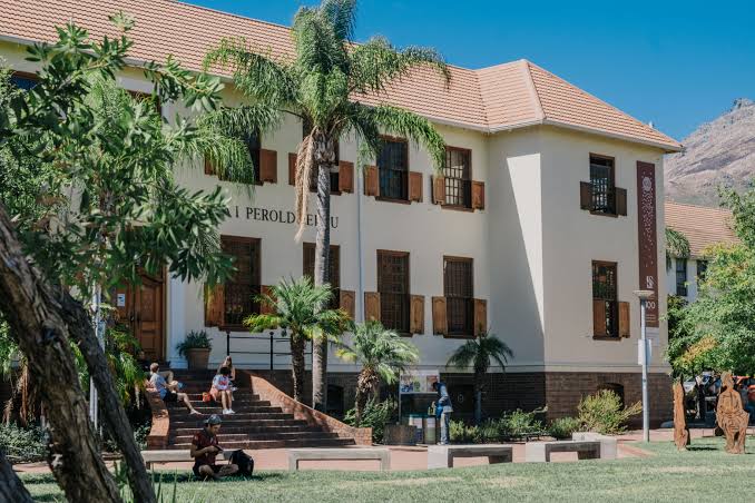 Stellenbosch University (campus scene): A sunny view of Stellenbosch University with a light building, palm trees, students relaxing outside, and a mountain in the background.