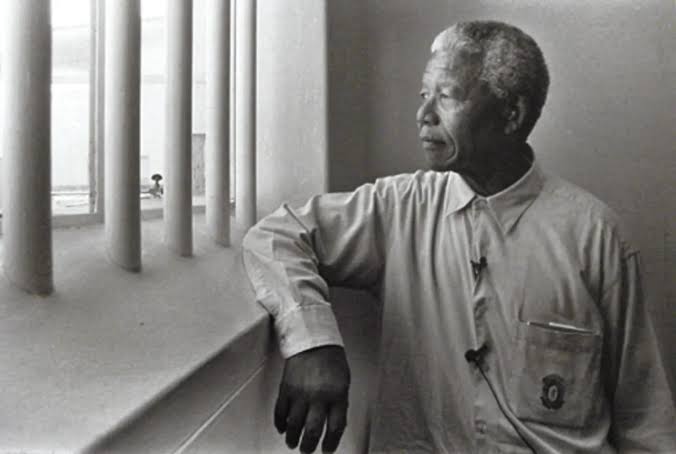 Nelson Mandela (Robben Island): A black and white photo of Nelson Mandela in a prison cell, looking thoughtfully out a barred window with one arm resting on the ledge.